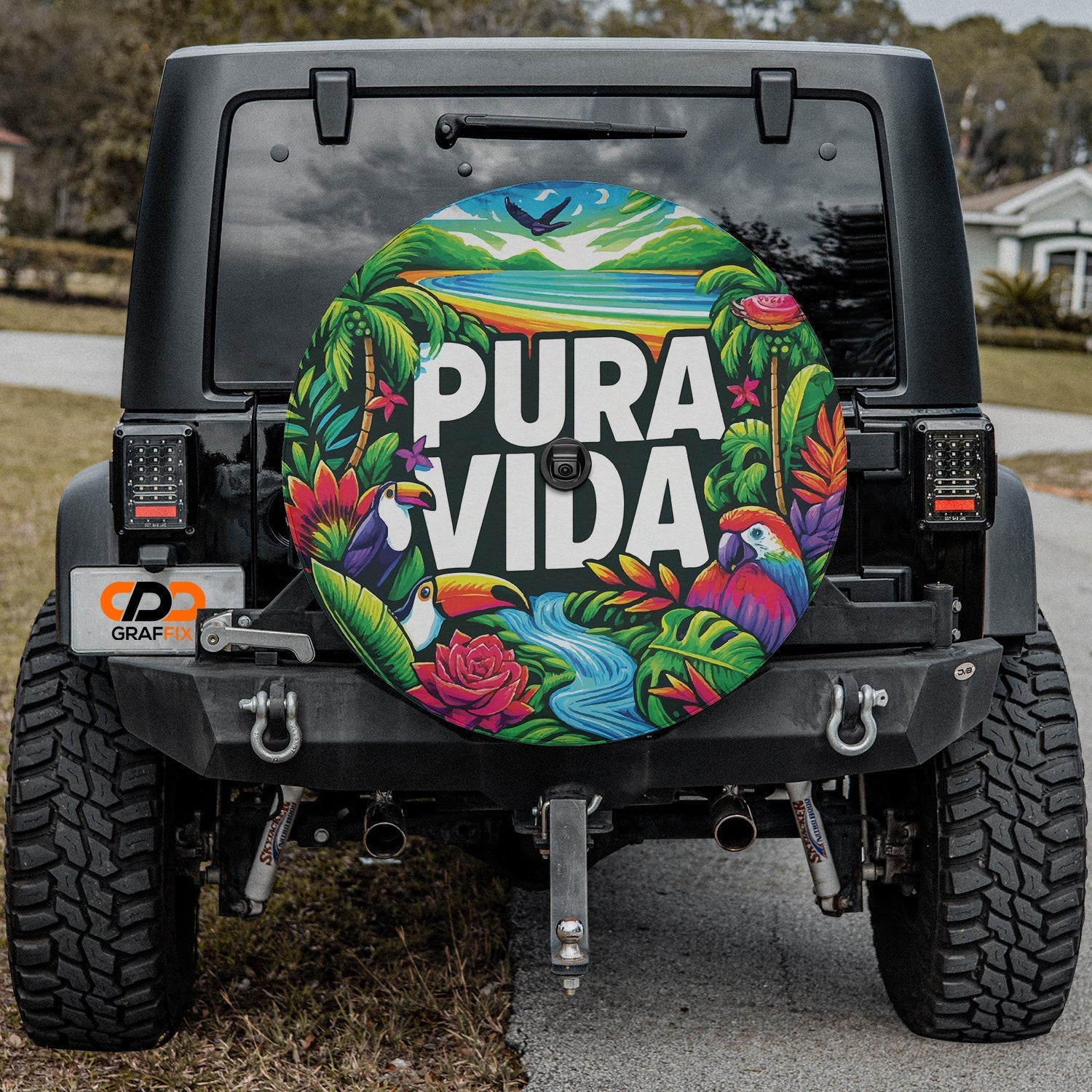 Pura Vida toucan macaw spare tire cover artwork mounted on black Jeep Wrangler in front yard scene