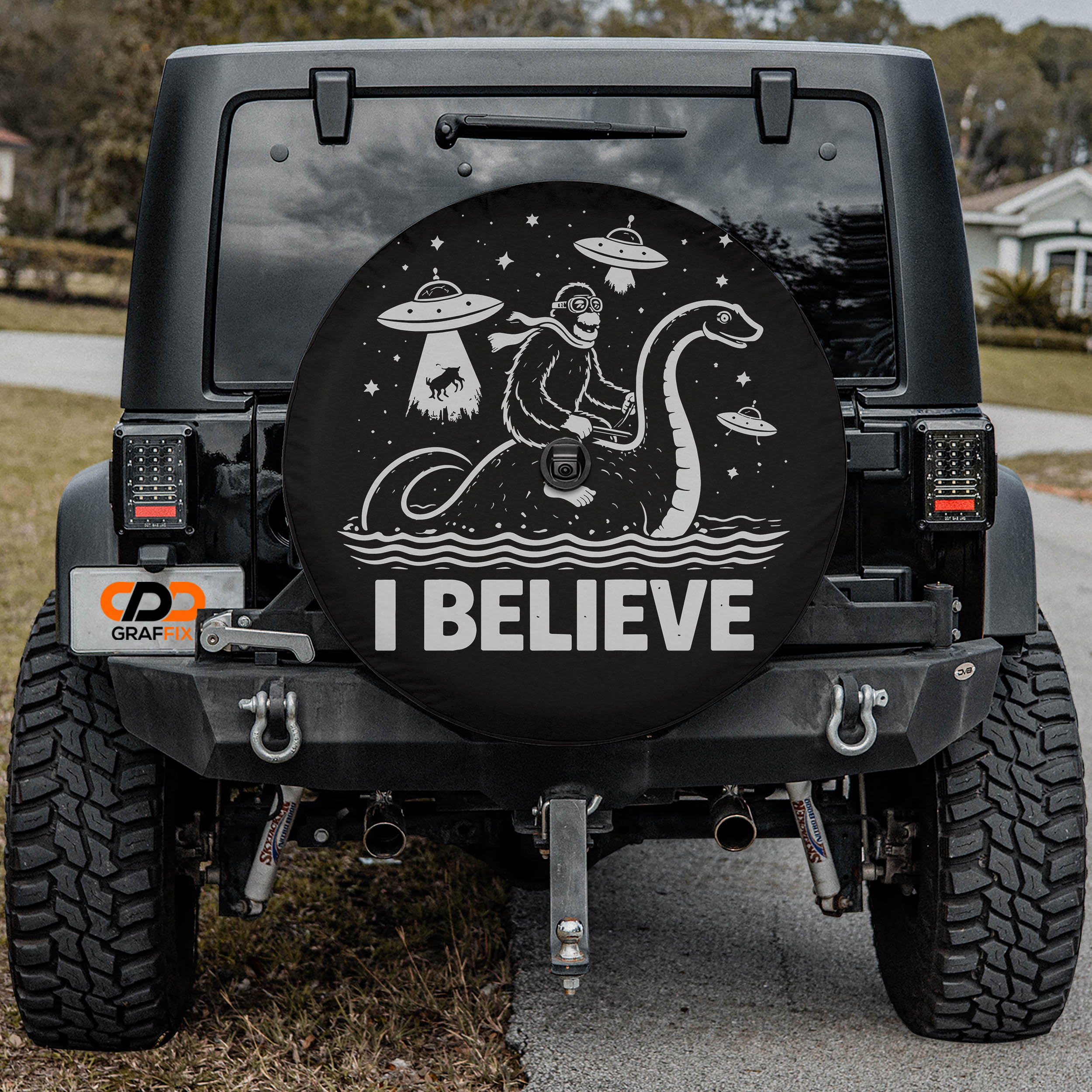 the back of a black Jeep Wrangler with a large, round spare tire cover on the rear of the vehicle. The cover features a cartoon image of a monkey riding a dinosaur on a boat, along with the text "I BELIEVE".