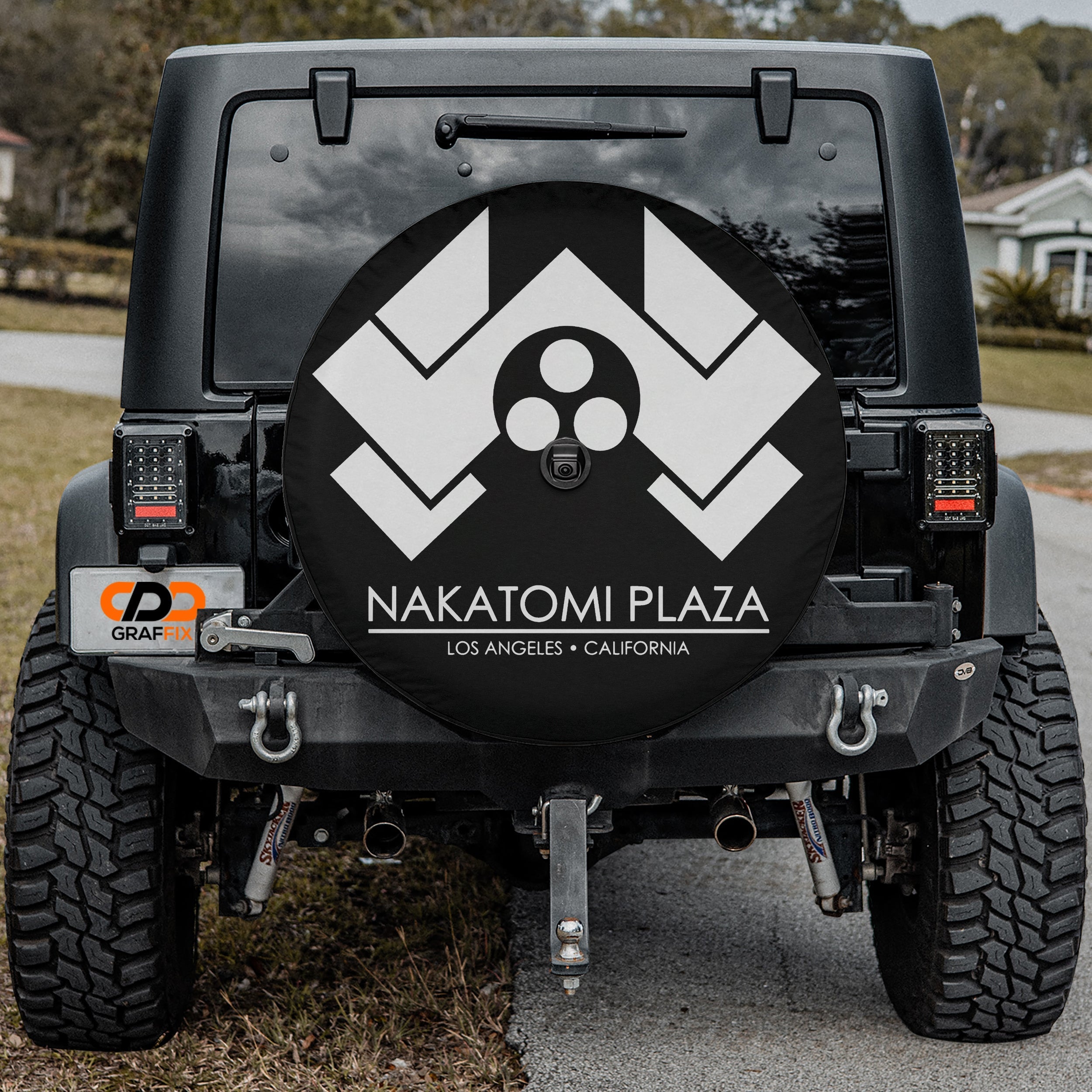 Nakatomi Plaza spare tire cover mounted on black Jeep Wrangler rear with white logo and camera cutout