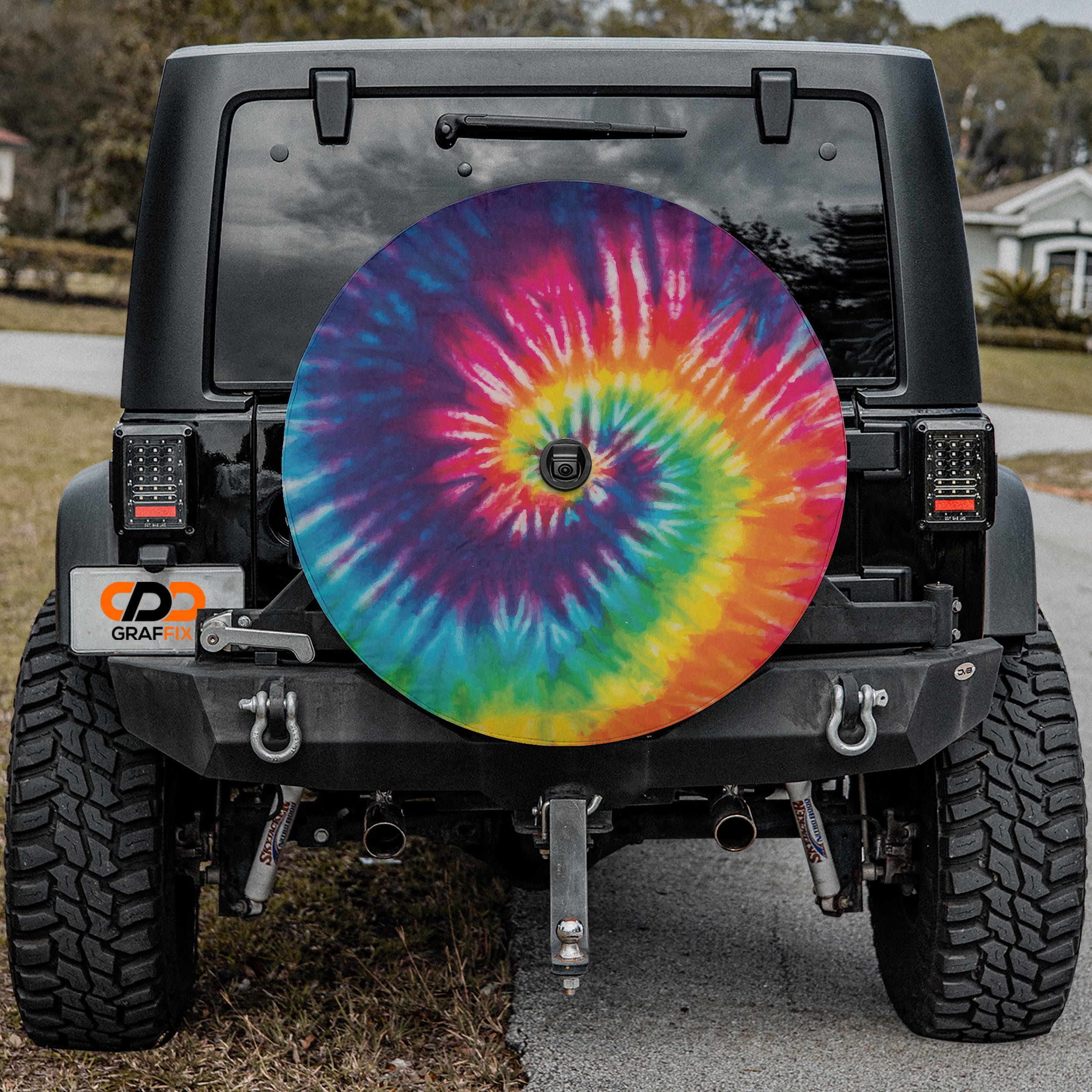 Bright tie-dye rainbow swirl tire cover on black Jeep Wrangler rear in driveway with houses behind