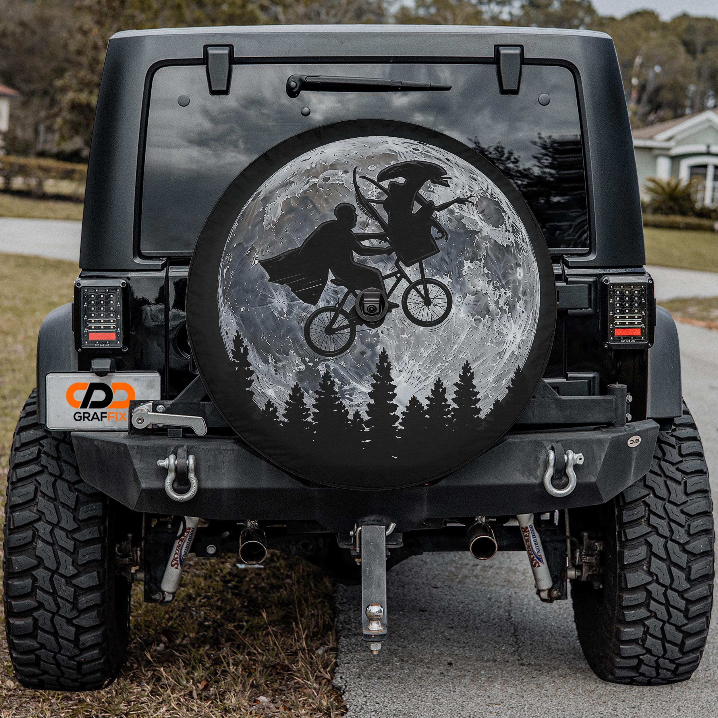 a black Jeep Wrangler with a large tire cover on the rear of the vehicle. The cover depicts a silhouetted image of two people riding a bicycle against a backdrop of a full moon and trees.