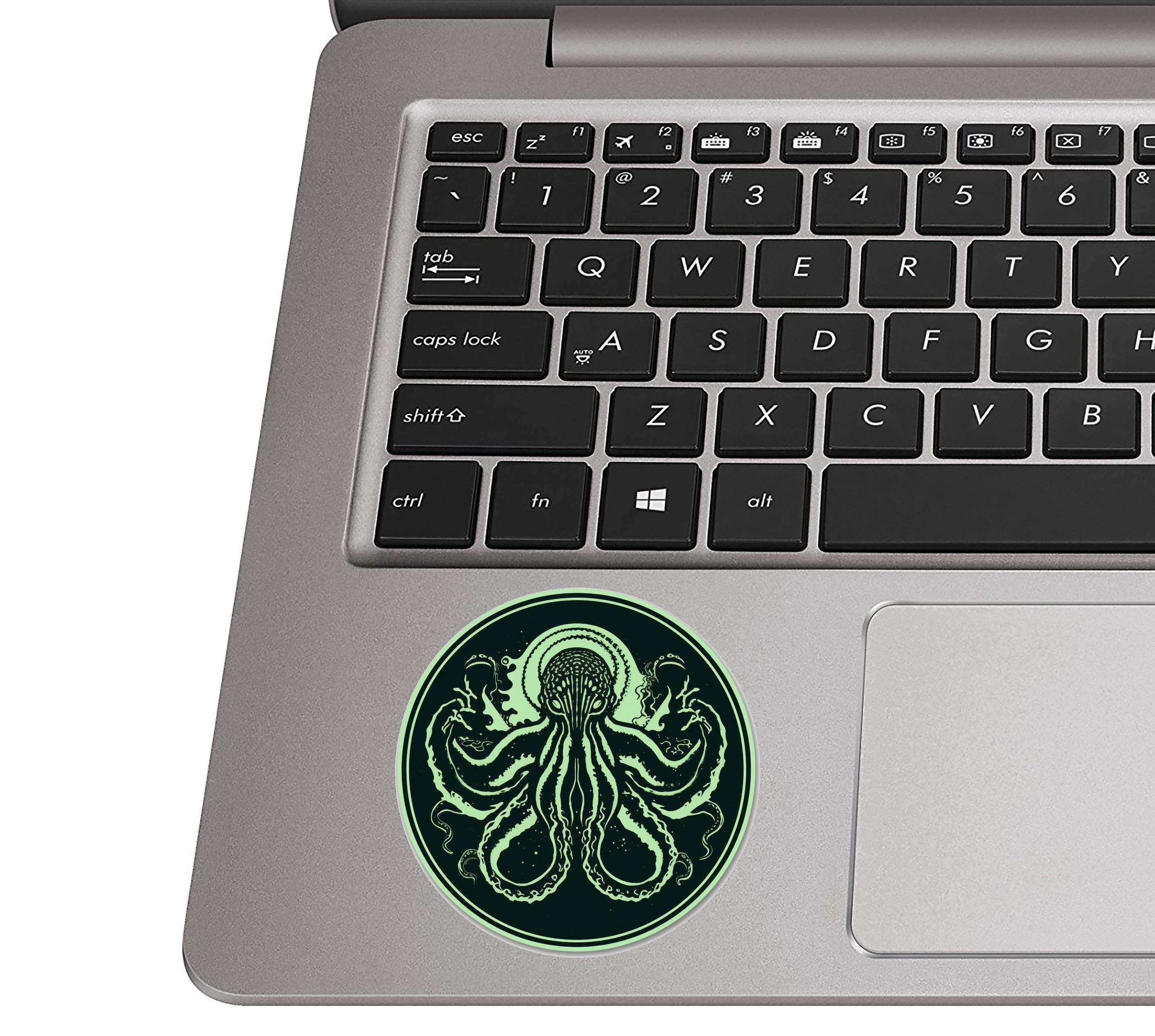 Minimalist Cthulhu green octopus circle sticker on silver laptop palm rest beside black QWERTY keys for scale